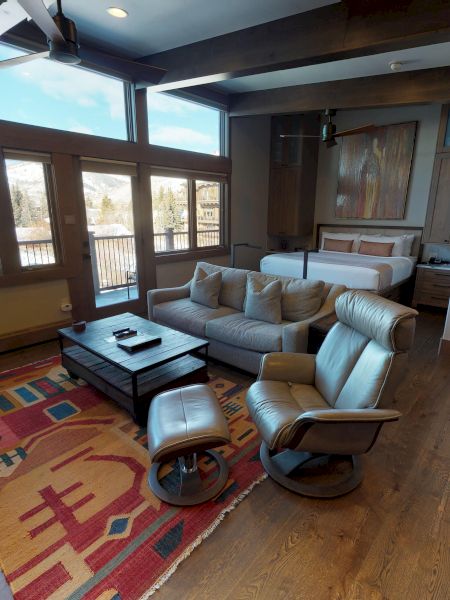 A cozy living space with large windows, modern furniture, and wooden flooring. Colorful rug and barstools accent the room's decor.