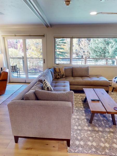 A bright living room with beige couches, a TV on a green cabinet, a stone fireplace, and large windows with balcony doors.