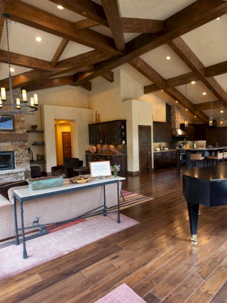 A rustic open-plan living room and kitchen with exposed beams, a stone fireplace, and a pool table in front.
