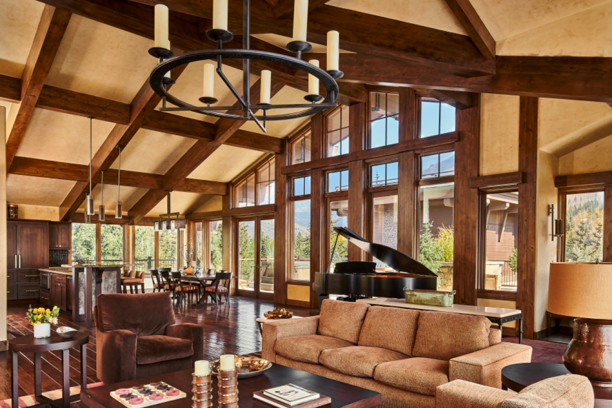 A spacious living room with a large window, chandelier, sofas, piano, and dining area, featuring wooden beams and furniture.