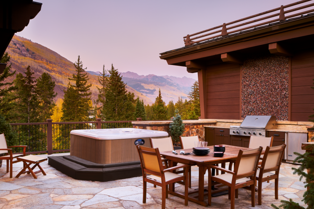 Outdoor patio with a hot tub, dining table, chairs, grill, and mountain view. The scene is set at sunset with surrounding trees.