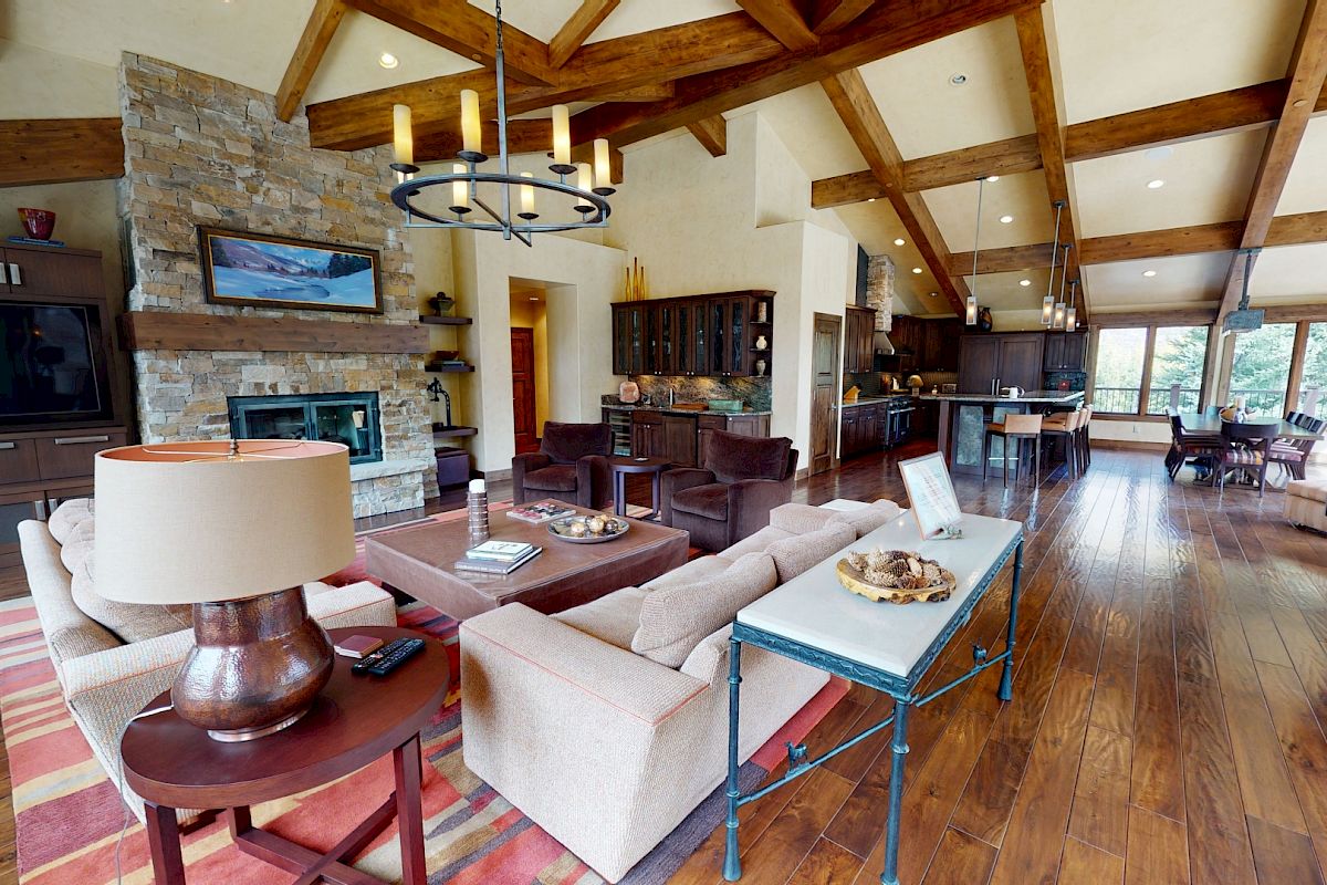 A spacious living area with wooden beams, stone fireplace, modern furniture, and a dining area leading to a kitchen, all well-lit.
