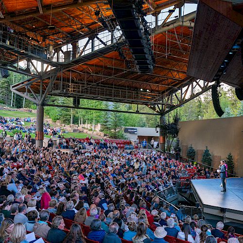 An outdoor amphitheater with a large audience and a speaker on stage, surrounded by a scenic, wooded area.