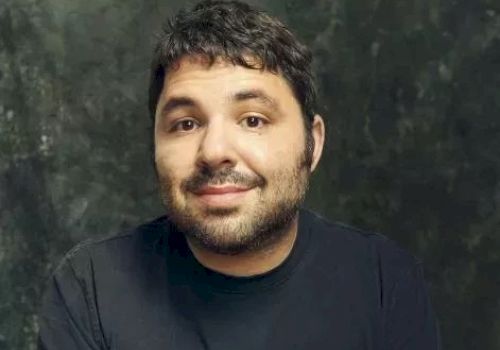 A man with short dark hair and a light beard, wearing a black shirt, smiles softly against a dark textured background.
