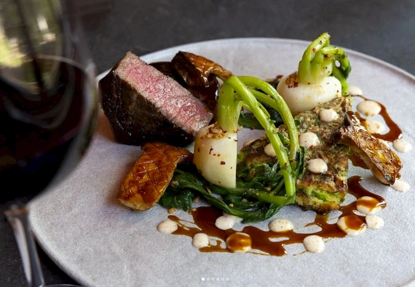 A plated fine-dining dish with a seared steak slice, greens, creamy swirls, and almond praline, all drizzled with sauces for a chic presentation.