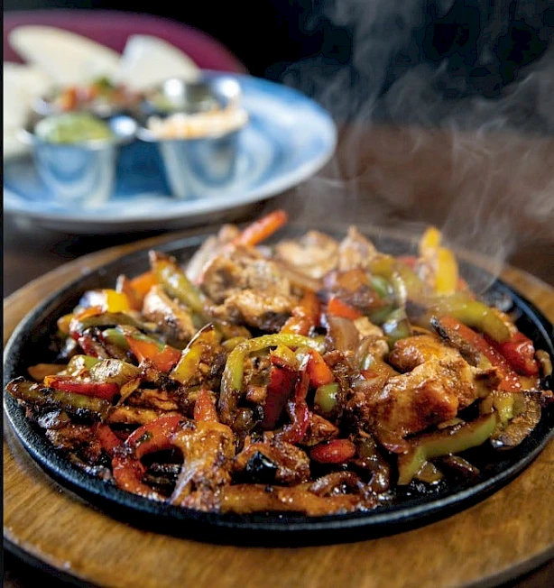 Sizzling sizzling fajita-style dish with peppers, onions, and meat on a hot skillet; steam rising, plate of toppings in the background.