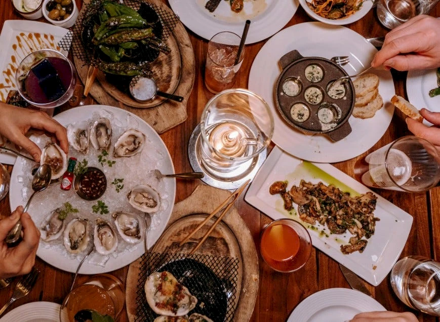 A lively dinner scene with various plates of oysters, scallops, and appetizers, drinks, candles, and people sharing a large table. End.