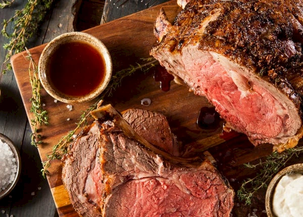 A sizzling roast beef on a cutting board, pink and juicy, with au jus dipping sauce and herbs for garnish on the side.