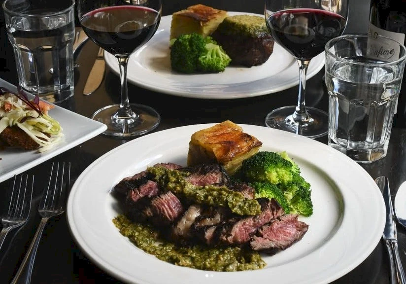 A gourmet steak dish with broccoli, potatoes, and a pesto-like sauce, plus side veggies and red wine glasses on a formal table.