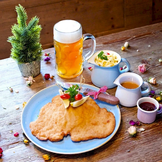 Beer, fried schnitzel on a blue plate with lemon and herbs, a mug of beer, a small pot of sauce, a tiny pot of gravy, and a teacup on a rustic wooden table with flowers and a small plant.