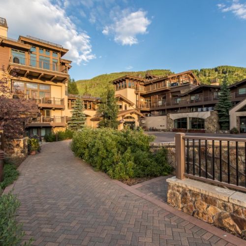 Cozy mountain resort with stone paths, brick buildings, balconies, manicured shrubs, and a lakefront railing against green hills under a blue sky.