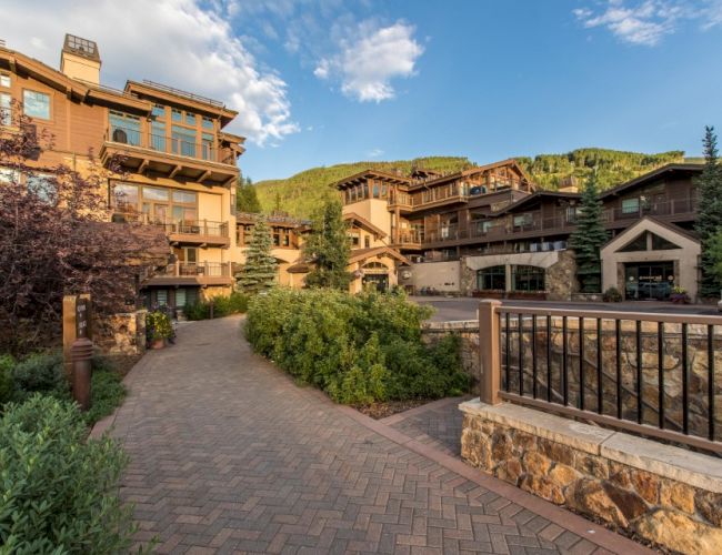 Cozy mountain resort with stone paths, brick buildings, balconies, manicured shrubs, and a lakefront railing against green hills under a blue sky.