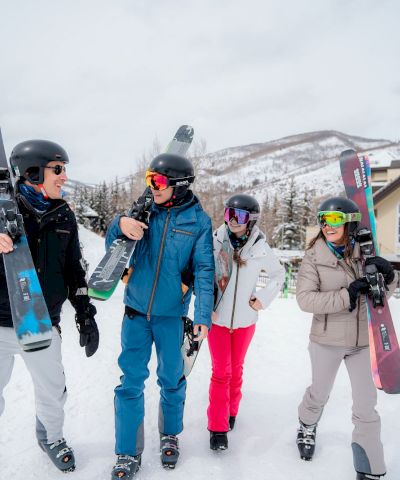 Four friends in snowsuits with skis and helmets stand on a snowy slope, posing for a photo at the base of Golden Peak