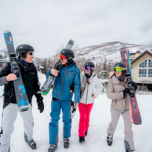 Four friends in snowsuits with skis and helmets stand on a snowy slope, posing for a photo near a chalet.