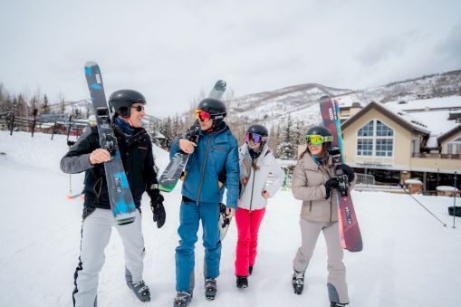 Four friends in snowsuits with skis and helmets stand on a snowy slope, posing for a photo at the base of Golden Peak