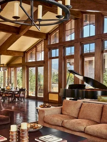 Luxurious living room with high vaulted ceilings, large wood beams, floor-to-ceiling windows, plush sofas, grand piano, and warm natural light.