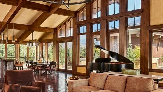 Luxurious living room with high vaulted ceilings, large wood beams, floor-to-ceiling windows, plush sofas, grand piano, and warm natural light.