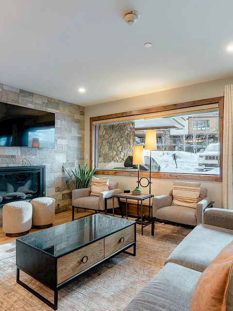 Cozy living room with a stone accent wall, mounted TV, gas fireplace, beige sectional sofa, glass coffee table, and large windows/doors letting in natural light.