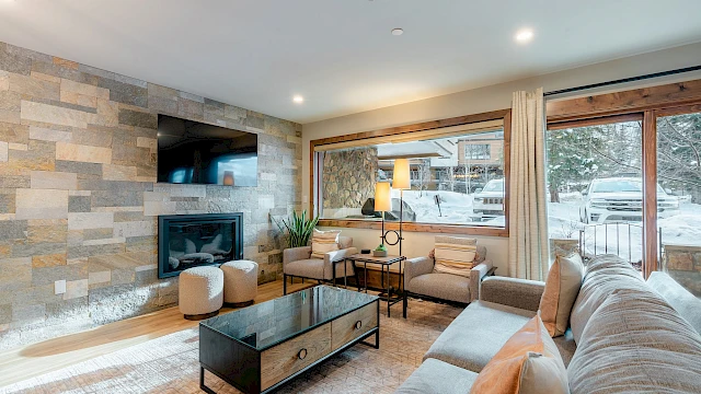 Cozy living room with a stone accent wall, mounted TV, gas fireplace, beige sectional sofa, glass coffee table, and large windows/doors letting in natural light.