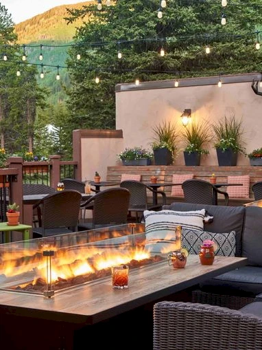 An outdoor lounge with wicker couches, a long table, string lights, fire pits, potted plants, and a deer head wall mount on a stucco backdrop.