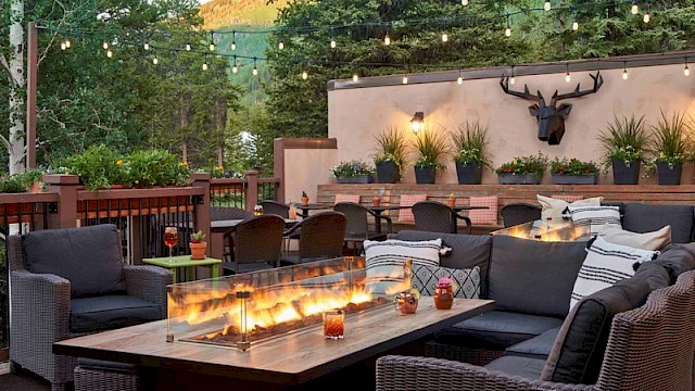 An outdoor lounge with wicker couches, a long table, string lights, fire pits, potted plants, and a deer head wall mount on a stucco backdrop.