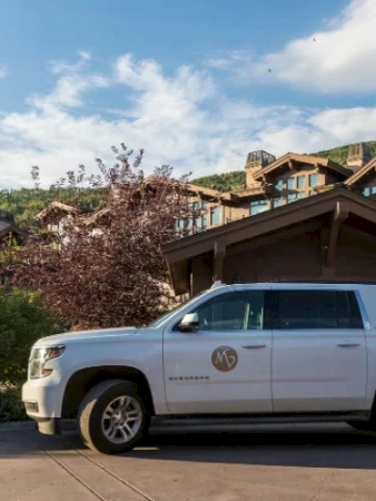 A white SUV parked in front of a resort-style building with balconies, mountains in the background, and a wooden entryway, under a blue sky.