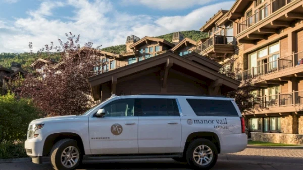 A white SUV parked in front of a resort-style building with balconies, mountains in the background, and a wooden entryway, under a blue sky.