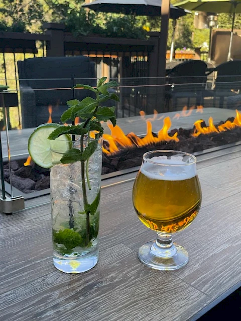 Two drinks on a wooden table near a fire pit: a tall mojito with mint and cucumber, and a small glass of beer, outdoor setting.