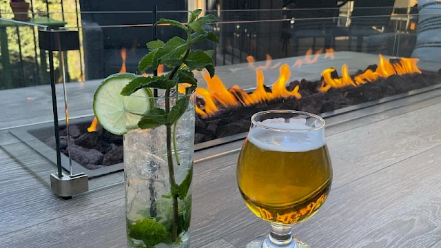 Two drinks on a wooden table near a fire pit: a tall mojito with mint and cucumber, and a small glass of beer, outdoor setting.