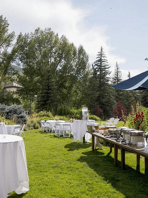 An outdoor garden party setup with white-clothed round tables, buffet stations under a blue umbrella, and lush green landscaping in a sunny yard.