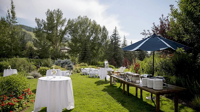 An outdoor garden party setup with white-clothed round tables, buffet stations under a blue umbrella, and lush green landscaping in a sunny yard.