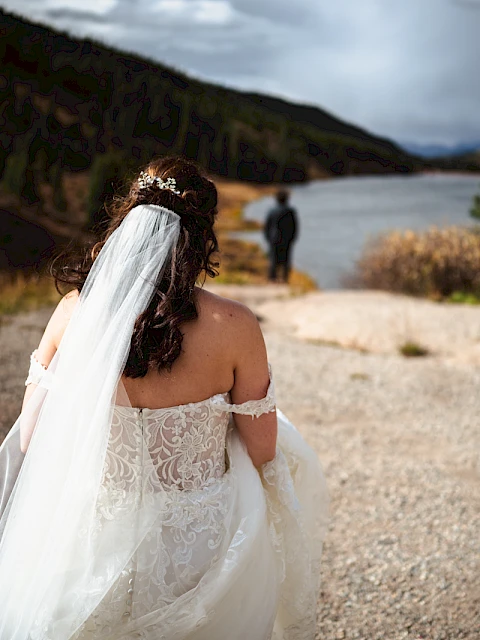 A bride in a lacy white wedding dress and veil walks along a rocky beach toward a man near the water, with hills in the background.