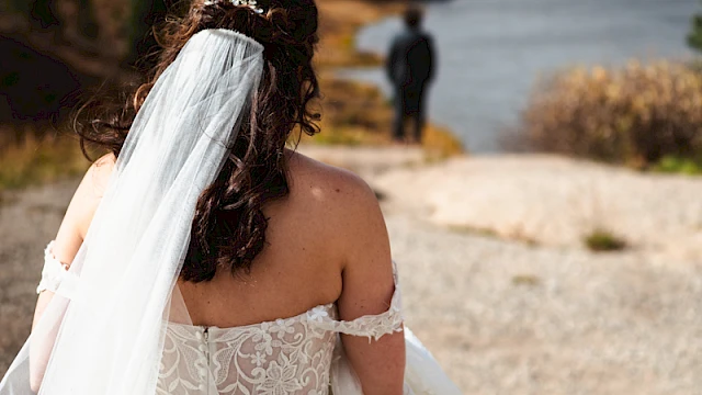 A bride in a lacy white wedding dress and veil walks along a rocky beach toward a man near the water, with hills in the background.