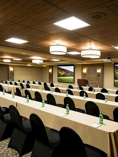 A conference room set for a meeting with long tables, black chairs, bottled water, and large screens displaying scenic images, lit by ceiling lights.