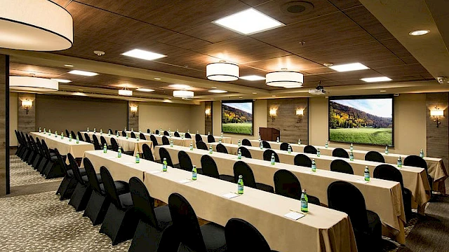 A conference room set for a meeting with long tables, black chairs, bottled water, and large screens displaying scenic images, lit by ceiling lights.