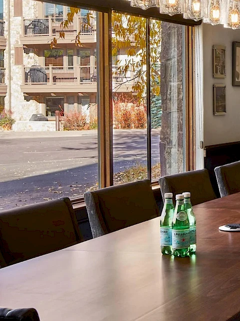 A long conference room with a wooden table, multiple chairs, bottled water, glass pendant lights, a wall-mounted TV, and large windows.