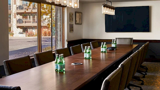 A long conference room with a wooden table, multiple chairs, bottled water, glass pendant lights, a wall-mounted TV, and large windows.