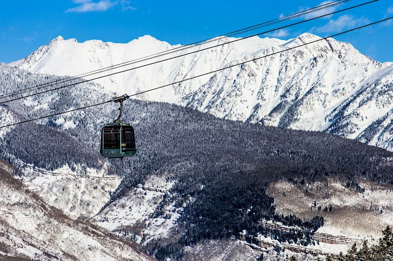 A snowy mountain landscape with a cable car suspended on wires, blue sky above and a dark forested slope below.