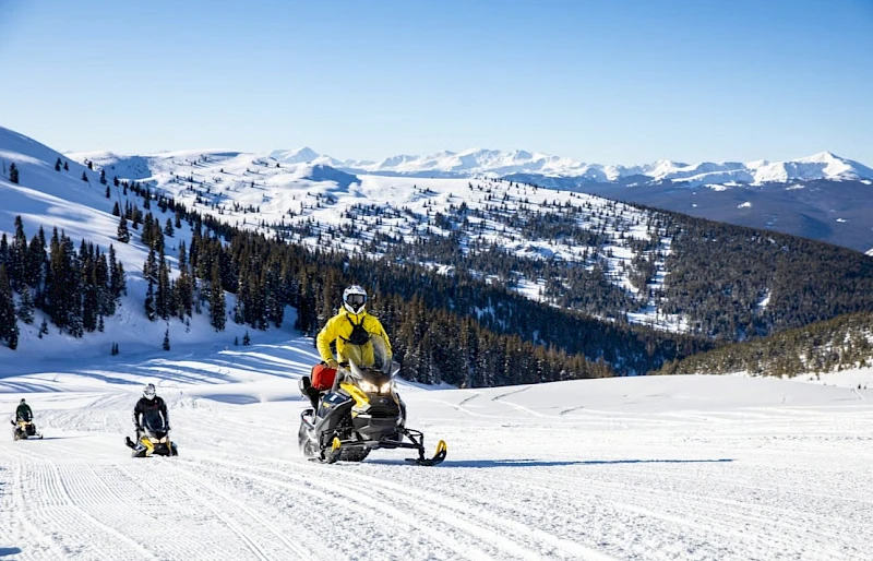 People snowmobiling on a snowy mountain slope with pine trees and distant peaks, clear blue sky, one rider in yellow ahead.