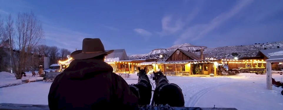 A snowy village at dusk viewed from a sleigh, with a person in a hat steering horses toward warmly lit buildings.
