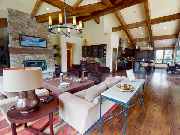 A spacious, modern living room with exposed wooden beams, a stone fireplace, and a mix of beige sofas, wood furniture, and a kitchen area in the background.