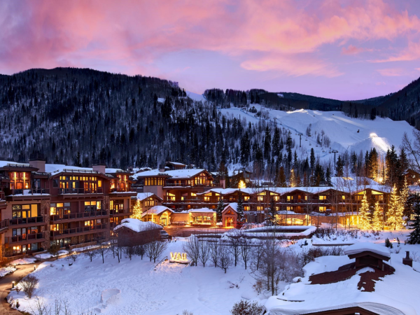 A snowy mountain village lit by warm lights at dusk, with ski slopes in the background and snow-covered trees and rooftops.