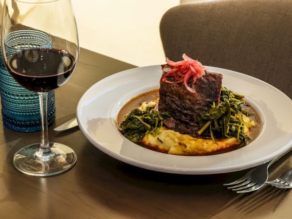 A plated steak with greens on a bed of vegetables and mashed potatoes, beside a glass of red wine.