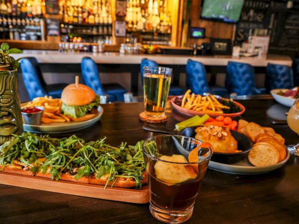 A bar scene with burgers, fries, wings, dips, and drinks on a wooden table; greenery garnish and assorted plates create a lively feast.