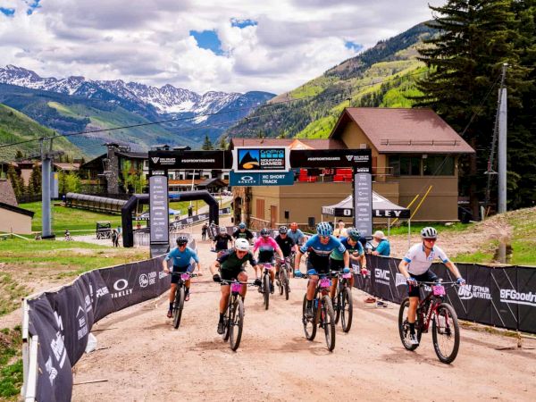 Cyclists race downhill on a dirt path near a mountain village, crossing a finish line under banners with alpine scenery and snow-capped peaks behind them.