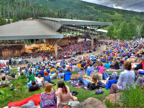 A large outdoor amphitheater filled with people watching a live performance on a covered stage in a green, hillside setting.