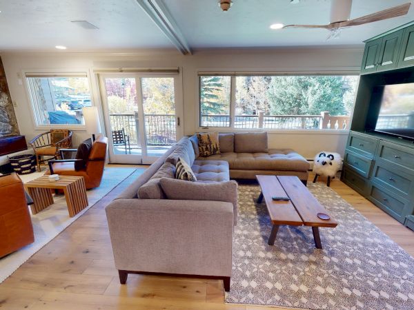Cozy living room with a green built-in cabinet wall, large sectional sofa, wooden coffee table, stone fireplace, and big windows overlooking a deck, all on a light wood floor.