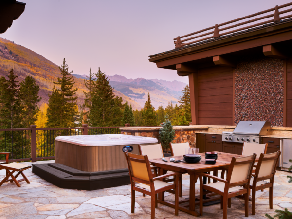 A mountain retreat patio with a hot tub, outdoor dining table, chairs, and a grill, overlooking pine trees and distant hills at sunset.