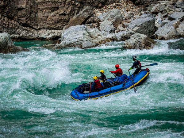 A blue inflatable raft with helmeted rafters shoots through turquoise whitewater rapids in a rocky canyon.