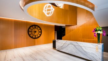 This image shows a modern lobby with a reception desk, a large winding staircase, a wall clock, and a vase of flowers on the desk.